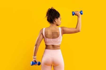 Téléchargez les photos : Vue arrière d'une puissante femme afro-américaine activement engagée dans une routine d'exercice de poids, mettant en valeur la puissance et la forme physique dans un cadre lumineux isolé - en image libre de droit