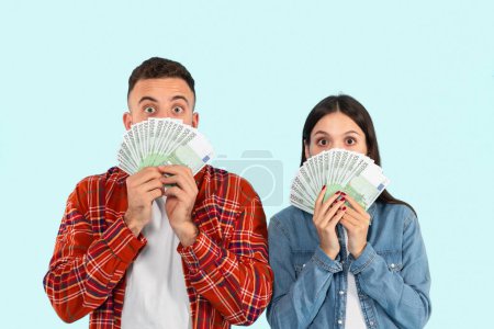 Un homme et une femme sont montrés cachant la moitié de leur visage avec de l'argent étalé, exprimant surprise et excitation. Ils se tiennent sur un fond bleu