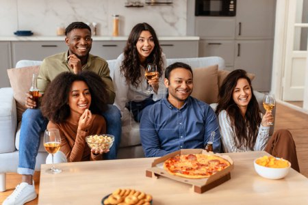 Diverse friends are gathered around a table enjoying pizza and wine, symbolizing camaraderie and leisure time at a cozy home