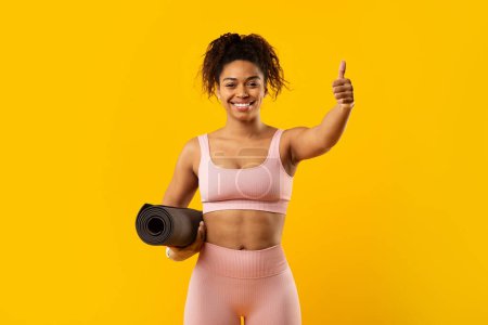 Cheerful african american woman in sportswear holds a yoga mat and gives a thumbs up against a vibrant yellow background