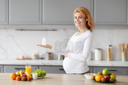 Téléchargez les photos : La femme européenne, visiblement en attente, présente une bouteille de compléments alimentaires, soulignant leur rôle dans la nutrition de la grossesse, dans une cuisine - en image libre de droit