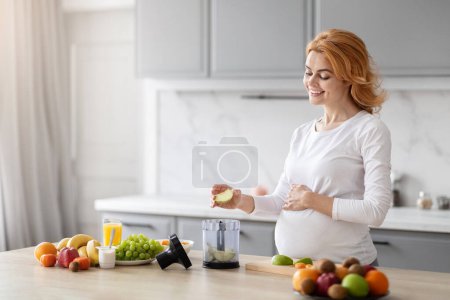 A cheerful pregnant woman is standing in a well-lit kitchen, preparing a healthy smoothie with fresh fruits and vegetables