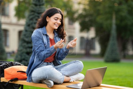 Photo for Positive young indian woman student have video call with classmate outdoors, looking at laptop screen, showing love gesture - Royalty Free Image