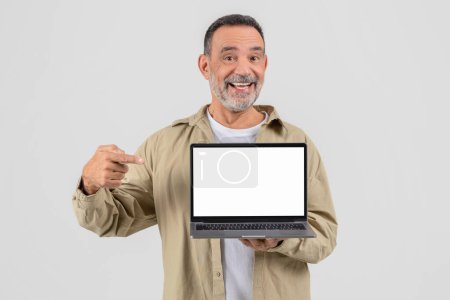 Téléchargez les photos : Homme mûr joyeux présentant un ordinateur portable avec un écran blanc, habillé de façon désinvolte, debout sur un fond gris - en image libre de droit
