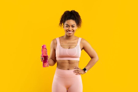Téléchargez les photos : Une jeune femme afro-américaine joyeuse en tenue de sport rose tient une bouteille d'eau réutilisable sur un fond jaune vif, prête pour une séance d'entraînement - en image libre de droit