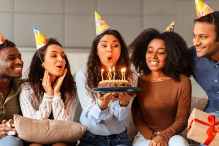 Photo for The moment of blowing out the birthday candles captured among multiracial young friends, marking a celebratory home event - Royalty Free Image