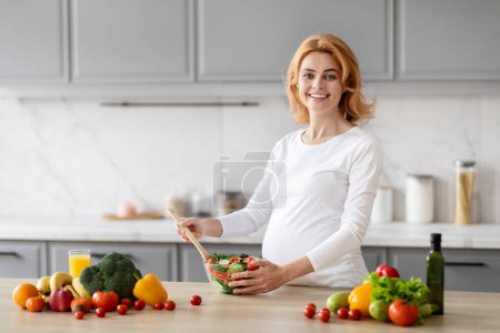 Téléchargez les photos : Une femme enceinte souriante dans un haut blanc préparant une salade saine avec des légumes frais dans une cuisine moderne lumineuse - en image libre de droit