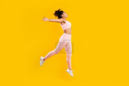 Téléchargez les photos : Image dynamique d'une femme afro-américaine affichant énergie et mouvement, mi-saut en tenue d'entraînement rose sur fond jaune vif - en image libre de droit