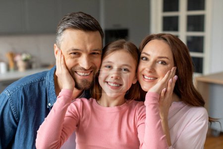 Téléchargez les photos : Une famille souriante avec une fille au milieu embrassant ses parents dans un cadre de cuisine moderne. Ils semblent connectés et joyeux - en image libre de droit