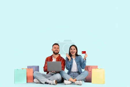Photo for A cheerful young man and woman sitting with legs crossed against a blue background, surrounded by colorful shopping bags, holding a credit card and a laptop - Royalty Free Image