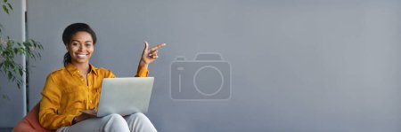 Foto de Feliz mujer de negocios negro sentado en la silla beanbag con ordenador portátil PC y señalando el dedo a un lado en el espacio libre, interior de la oficina. Lady Mostrando espacio de copia para su anuncio o texto, banner web - Imagen libre de derechos