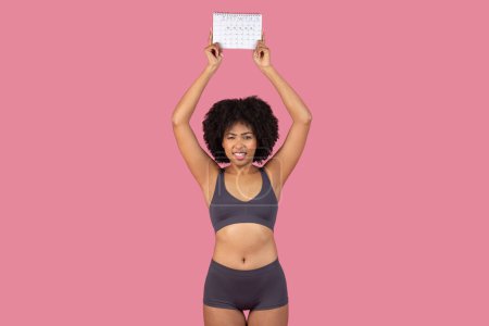 Photo for A black, sporty lady is isolated against a pink backdrop, energetically holding period calendar above her head with a concerned expression - Royalty Free Image
