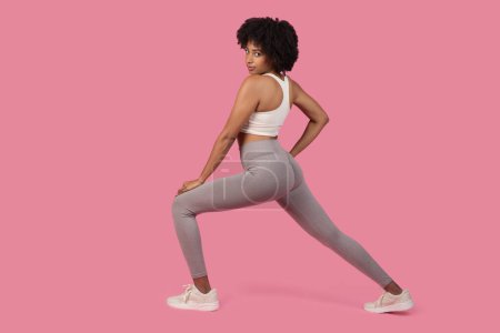 Téléchargez les photos : Une femme afro-américaine concentrée en leggings gris s'étirant avant l'exercice, faisant preuve de détermination, isolée sur rose - en image libre de droit