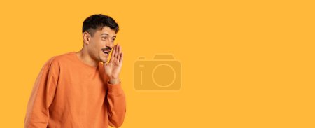 Photo for Man in an orange sweatshirt appears to whisper or speak secretly against a vivid orange background, with copy space - Royalty Free Image