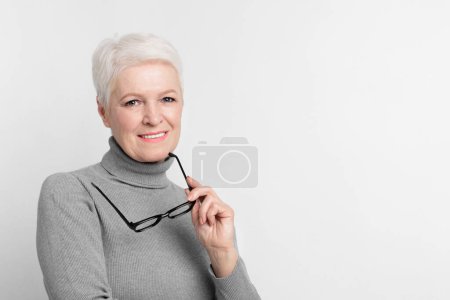 Elderly European woman holding glasses on grey background, showcasing s3niorlife sophistication, copy space