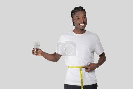 Téléchargez les photos : A fit young African American man with braided hair smiles as he holds a dumbbell in one hand and a measuring tape around his waist, suggesting health and exercise - en image libre de droit