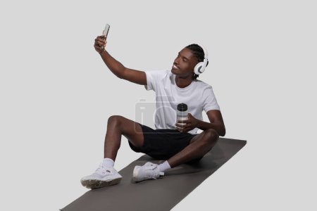 Photo for An african american guy with headphones is taking a selfie, sitting cross-legged on a gym mat isolated on a white background - Royalty Free Image