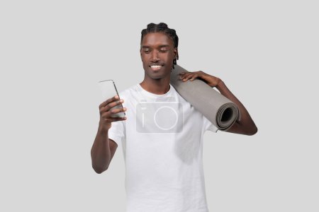Foto de Un hombre negro feliz con una camiseta blanca lleva una esterilla de yoga en el hombro mientras mira su teléfono móvil, sobre un fondo gris claro - Imagen libre de derechos