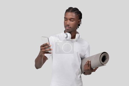 Ein junger afroamerikanischer Mann hält ein Smartphone in der Hand und trägt eine Yogamatte, was auf einen Lebensstil hindeutet, der Technologie und Fitness kombiniert