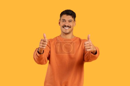 A millennial man with a moustache and a funny expression gives two thumbs up in a radiant orange sweater, isolated against an orange backdrop