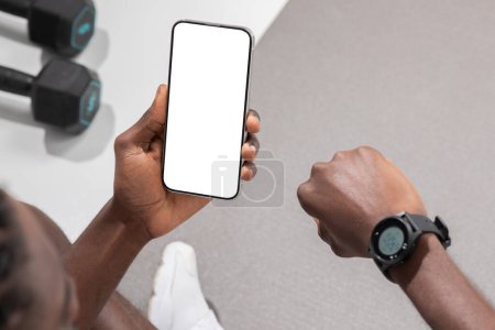 Photo for An african american athletic guy reviews his workout progress on a smartwatch and phone, representing sport dedication, isolated on a light backdrop - Royalty Free Image