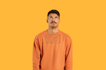 Photo for A millennial guy with a moustache looks up with a puzzled expression against a funny, isolated orange background - Royalty Free Image