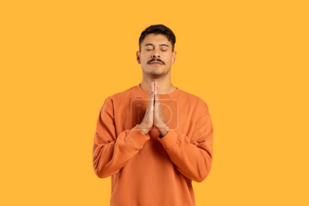 Photo for A serene man in an orange shirt stands with closed eyes and hands in prayer position against a yellow background - Royalty Free Image