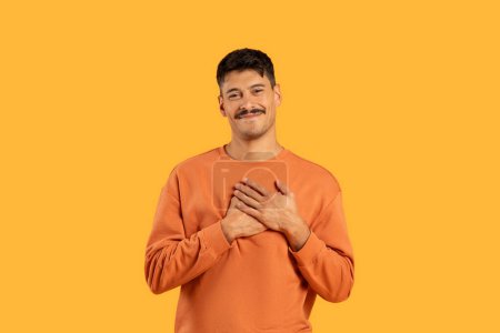 A candid portrait of a cheerful young man in an orange shirt, hands on heart, against a vibrant yellow background