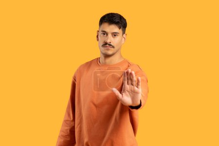 A man in an orange sweater stands against an orange backdrop, extending his hand forward in a stopping gesture