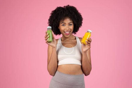 Foto de Deportiva dama afroamericana sosteniendo botellas de batido de desintoxicación verde y amarillo, promoviendo una alimentación saludable, aislada en rosa - Imagen libre de derechos