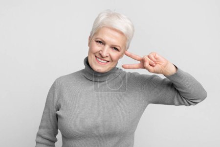 Téléchargez les photos : Gaie senior européen femme clignote un signe de paix près de son oeil, dépeignant un s3niorlife ludique moment - en image libre de droit