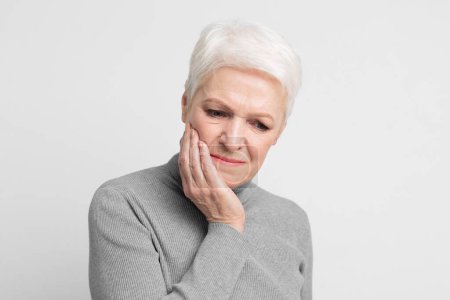 Téléchargez les photos : Une femme âgée européenne aux cheveux blancs courts en pull gris tient sa joue, montrant un inconfort ou une douleur dentaire, une scène pertinente pour s3niorlife - en image libre de droit