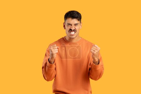 Ein junger Mann zeigt eine starke Geste der Verärgerung oder Wut und ballt seine Fäuste fest auf einem schlichten orangefarbenen Hintergrund
