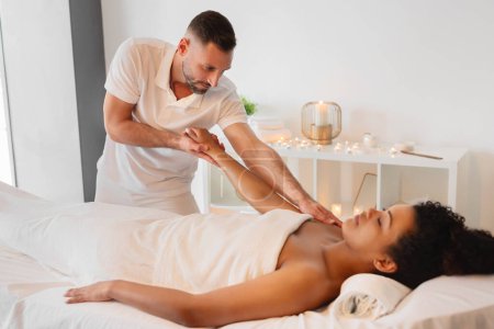 Téléchargez les photos : Un masseur qualifié offre un massage corporel thérapeutique à une femme afro-américaine détendue dans un cadre de spa serein, entouré de bougies et de lumière douce - en image libre de droit