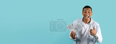 Excited man displaying a handful of cash with a thumbs up next to generous blank space on blue mockup background