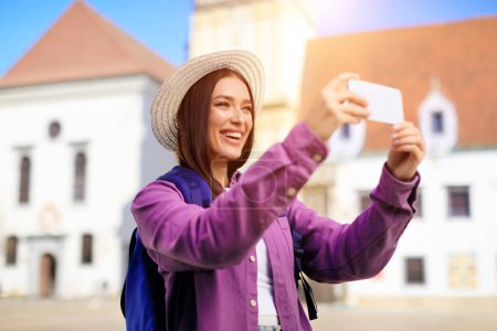 Téléchargez les photos : Femme souriante avec chapeau de soleil prenant un selfie avec son téléphone avec des bâtiments historiques derrière, concept de tourisme - en image libre de droit