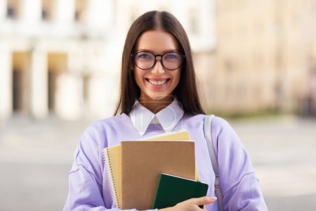 A cheerful young woman in glasses is holding textbooks, likely a student, with a blurred city background, radiating positivity and confidence