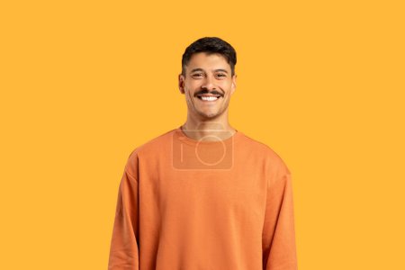 A millennial guy with a moustache smiling at camera, posing on a funny and vibrant isolated orange background