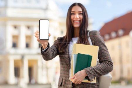 Téléchargez les photos : Joyeux jeune étudiante montre son écran de smartphone, vide pour la maquette, avec des cahiers à la main, espace de copie - en image libre de droit