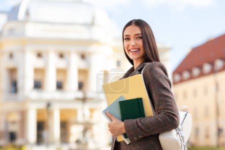 A young woman student in a plaid jacket with books smiles confidently, old architecture in background