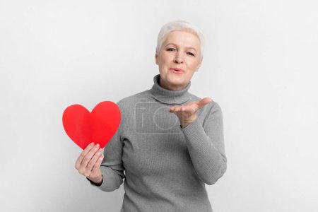 Téléchargez les photos : Femme européenne âgée aux cheveux courts tenant un cœur sur fond gris, symbolisant l'amour dans l'imagerie s3niorlife - en image libre de droit