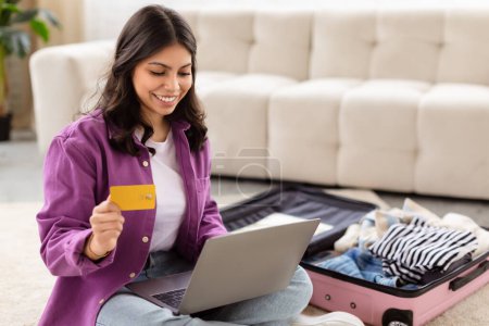 Young middle eastern woman in casual wear using a credit card and laptop to plan a trip, luggage open in front of her
