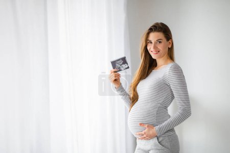 Téléchargez les photos : Une femme enceinte en robe rayée sourit tout en tenant une image échographique de son enfant à naître - en image libre de droit