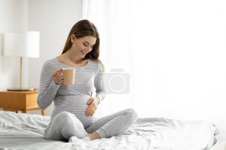 A smiling pregnant woman is relaxing in bed, holding a cup with both hands, and looking at her belly in a warmly lit room