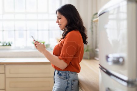 Foto de Una mujer de Oriente Medio en casa, utilizando su teléfono inteligente en una cocina bien iluminada y moderna, que encarna la conexión y la tecnología - Imagen libre de derechos
