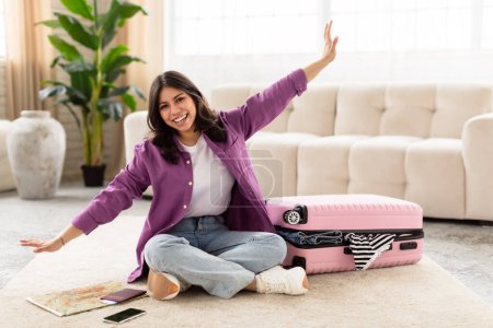 Photo for A joyous middle eastern woman with arms wide open sitting on the floor next to a pink suitcase, expressing travel excitement - Royalty Free Image