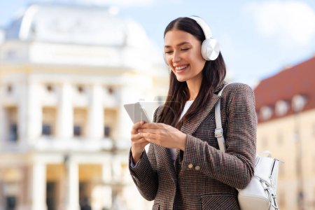 Téléchargez les photos : Une jeune femme en vêtements élégants sourit en utilisant son smartphone et en portant des écouteurs blancs dans un cadre urbain - en image libre de droit