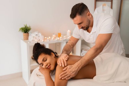 Téléchargez les photos : Masseur qualifié offrant un massage des bras dans un spa, avec une femme afro-américaine appréciant le traitement - en image libre de droit