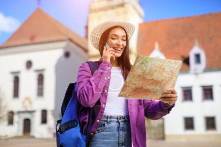 Téléchargez les photos : Femme souriante en tenue de voyage parlant au téléphone tout en tenant une carte, bâtiments historiques en arrière-plan - en image libre de droit