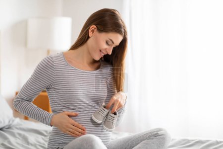Photo for European lady feeling her baby bump and holding baby shoes, evoking the warmth and expectancy of bringing a new life into her home during pregnancy - Royalty Free Image